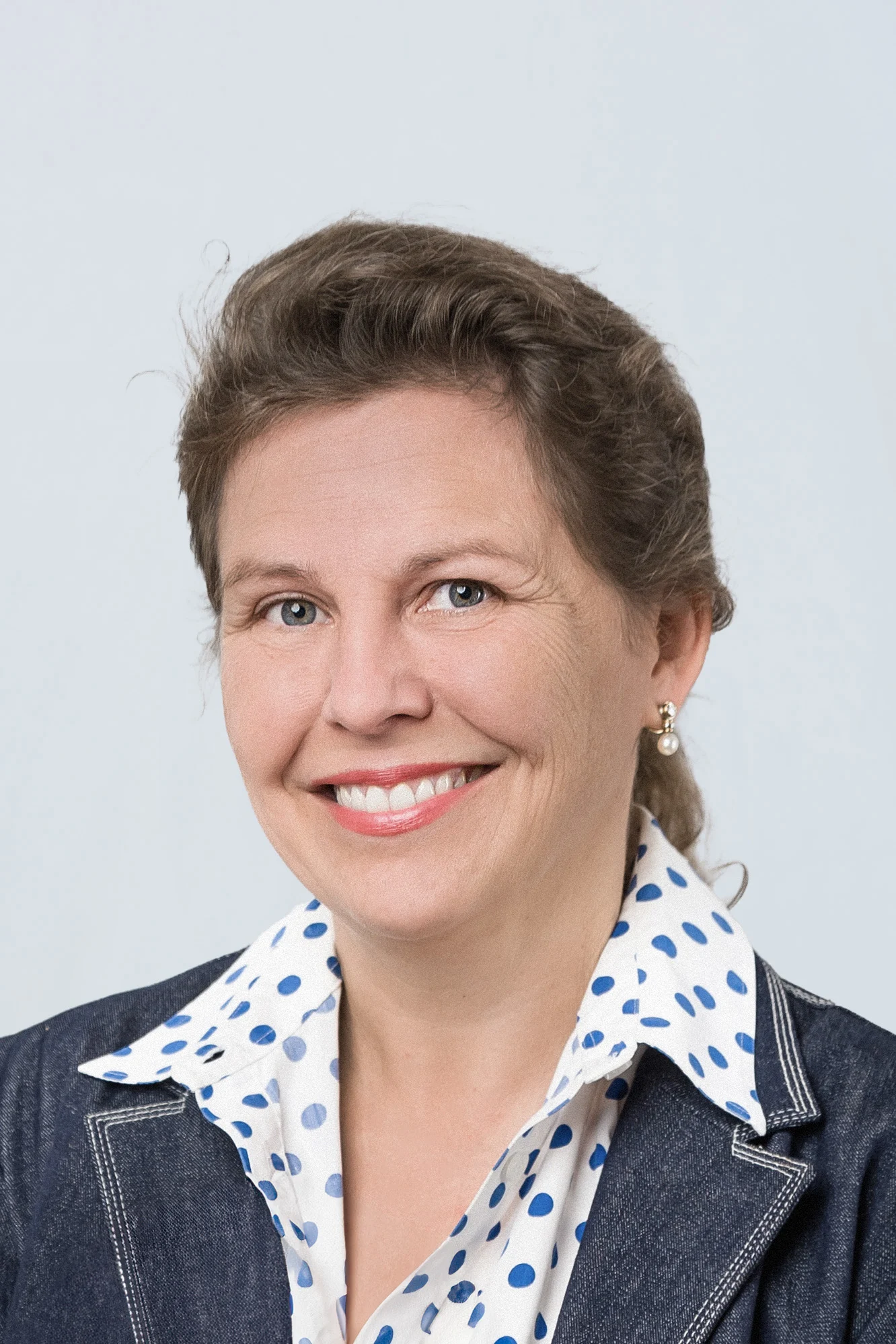 Porträt einer Frau mit braunen Haaren, die eine blaue Jacke und ein gepunktetes Hemd trägt.