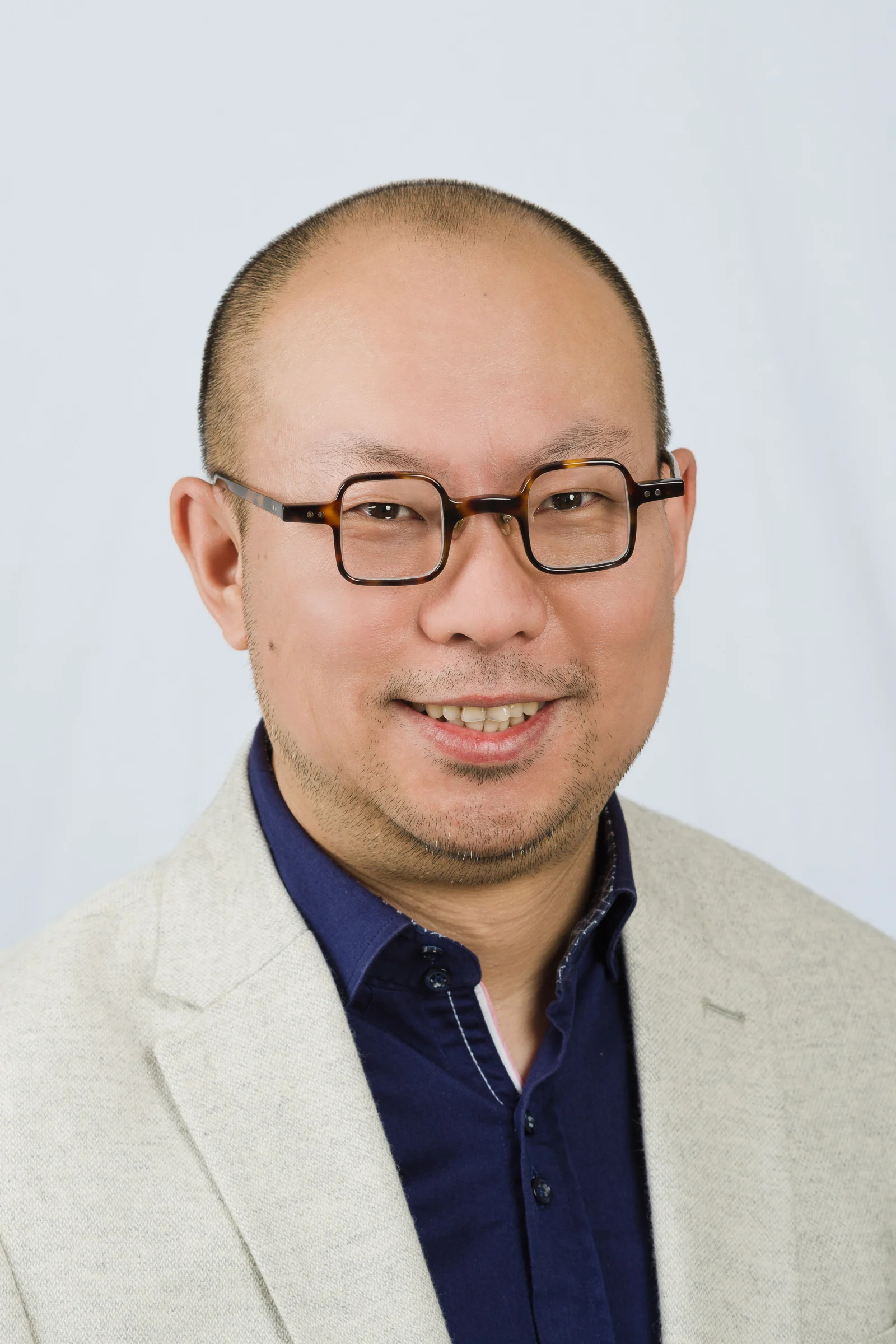 Porträt eines Mannes mit Glatze, Brille und hellgrauer Anzugjacke vor einem neutralen Hintergrund.