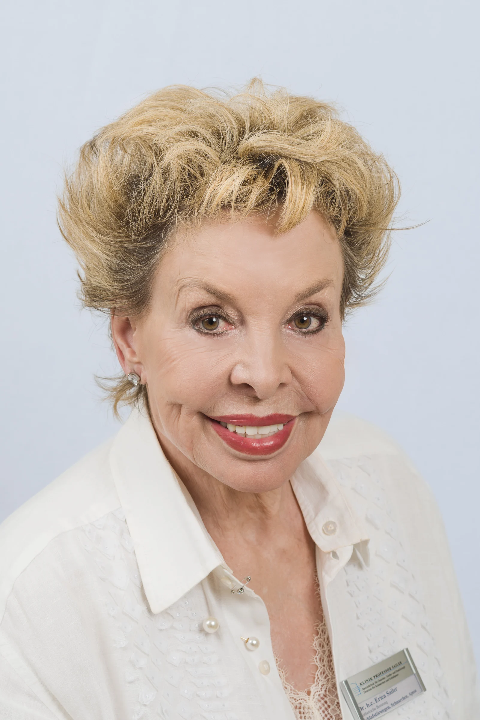 Portrait einer älteren Frau mit blonden, lockigen Haaren und einem weißen Oberteil.