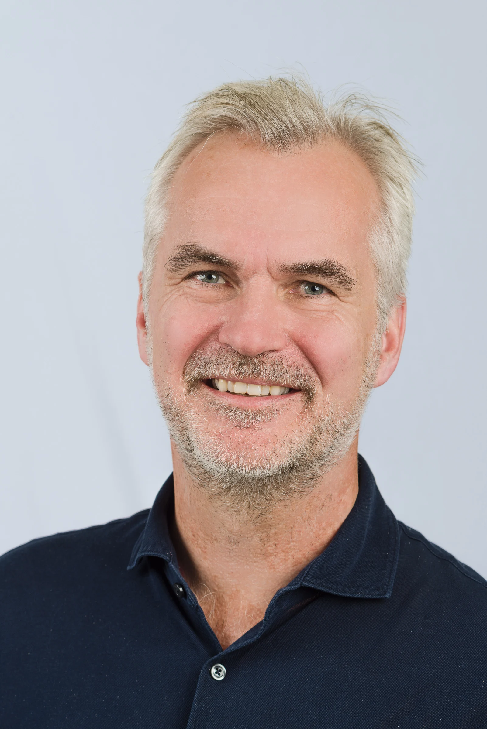 Porträt eines lächelnden Mannes mit grauen Haaren und Bart, in einem blauen Hemd.