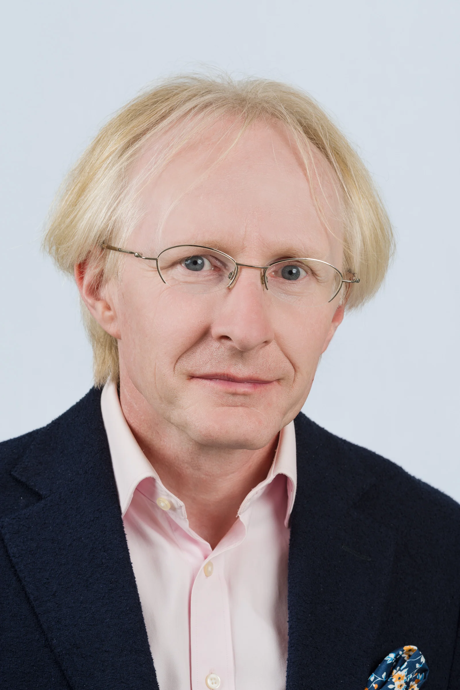 Porträt eines Mannes mit blonden Haaren, Brille und dunkler Jacke auf einem hellen Hintergrund.